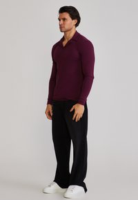 Bărbat în picioare purtând o cămașă lungă cu mâneci lungi de culoare mov închis, pantaloni negri largi și adidași albi, pe un fundal simplu, deschis.