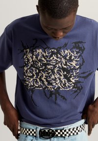 Navyfarbene Baumwoll-T-Shirt mit unregelmäßigem schwarz-braunem abstraktem Druck. Weiß-schwarz karierter Gürtel mit runder Schnalle, getragen mit hellen Jeans.