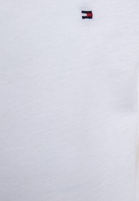 T-shirt in cotone bianco con una superficie liscia e un piccolo logo ricamato nei colori rosso, bianco e blu posizionato sul petto.