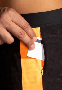 Svarta träningsshorts har en ljusorange meshficka med dragkedja, medan en hand stoppar in ett kort. Fickan har en kontrasterande textur.
