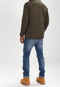Veste zippée vert olive avec col montant, arborant un patch sur l'épaule gauche, portée avec un jean bleu et des bottes marron clair.