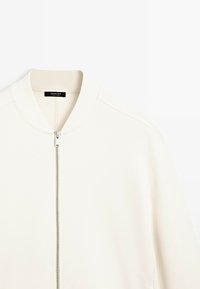 Veste bomber blanche avec un col côtelé et une fermeture éclair sur le devant. Fabriquée en tissu lisse, avec des coutures d'épaules cousues et sans motifs visibles.