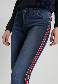 Mörkblå denimjeans med smal passform, prydda med röda och vita sidränder samt en metallknappvidaring i midjan. Slät textur.