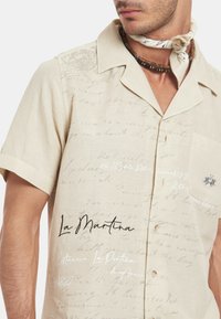 Kratká béžová lněná košile s krátkými rukávy, která má potisk ručně psaného textu a náprsní kapsu. Mezi výrazné detaily patří logo a dekorativní šití.