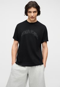 Νεαρός με μεσαίου μήκους σκούρα μαλλιά φοράει μαύρο μπλουζάκι με σχέδιο και ανοιχτό γκρι σορτς, στέκεται με τα χέρια στις τσέπες.