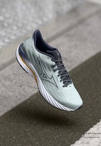 Hellgrau Laufschuh mit dunkelgrauen Schnürsenkeln und Akzenten, weiße Sohle mit orangefarbenem Streifen, in der Luft über einer Asphaltoberfläche eingefangen.