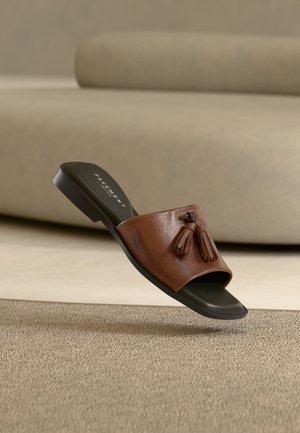 Braune Ledersandale zum Hineinschlüpfen mit Quasten auf schwarzer Sohle, die über einem beigen Teppich in einem neutralen Innenraum schwebt.