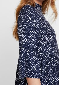 Granatowa sukienka z białym wzorem liści, krótki rozkloszowany rękaw, wysoka dekolt i marszczona talia, wykonana z lekkiego materiału.