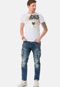 Weißes kurzärmliges T-Shirt mit schwarzem und goldenem Grafikdesign, kombiniert mit lässigen, ausgebleichten blauen Cargo-Jeans, die mehrere Taschen und Reißverschlüsse aufweisen.