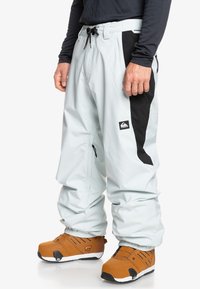 Pantalones de nieve azul claro con paneles laterales negros, cinturilla ajustable y cremalleras en los bolsillos, combinados con botas marrones con detalles en negro.