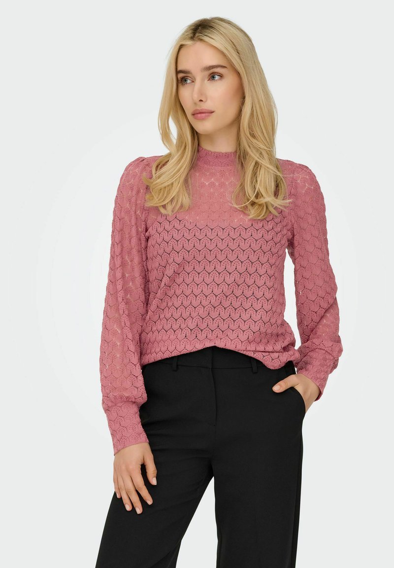 Femme blonde aux cheveux détachés, portant un chemisier en dentelle rose à manches longues et un pantalon noir, debout avec une main dans la poche, regardant vers la droite.