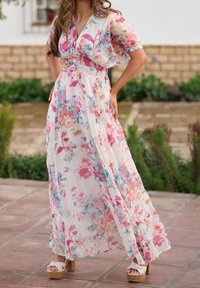 Femme portant une robe maxi blanche à fleurs avec manches courtes et sandales à plateforme, debout sur une terrasse carrelée avec des plantes en arrière-plan.