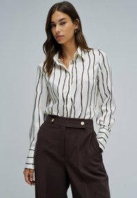 Blusa blanca con rayas verticales marrones, con botones en la parte delantera y cuello. Combinada con pantalones marrones de cintura alta con detalles dorados.