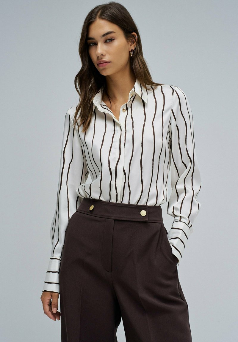 Blusa blanca con rayas verticales marrones, con botones en la parte delantera y cuello. Combinada con pantalones marrones de cintura alta con detalles dorados.