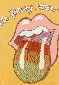 Der gelbe Stoff zeigt ein buntes, mit Pailletten versehenes Logo der Rolling Stones, mit einem auffälligen Zungendesign in Pink, Orange und Grün.