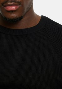 Schwarzer Rippstrickpullover mit einem glatten runden Ausschnitt und Raglanärmeln, der ein strukturiertes Muster auf einer schmalen Silhouette zeigt.