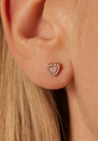 Ohrringe in Herzform aus goldfarbenem Metall mit pinkfarbener Einlage und umgebenden klaren Edelsteinen, die eine strukturierte Oberfläche auf der Haut zeigen.