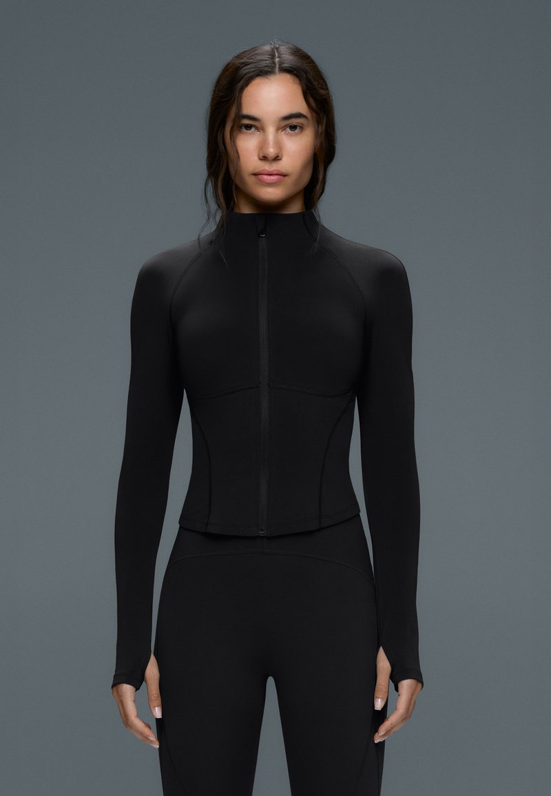 Femme aux longs cheveux foncés porte une veste noire ajustée avec fermeture éclair à l'avant et un legging assorti, debout devant un fond gris uni.