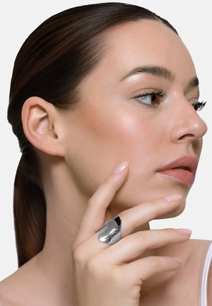 Kobieta o brązowych włosach związanych nisko w kitkę, dotykająca podbródka, nosząca duży, błyszczący, srebrny pierścionek na palcu wskazującym.