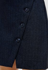 Marineblaue Wickelrock aus strukturiertem Stoff, der einen diagonal geschnittenen Schnitt aufweist und zwei dunkle Kunststoffknöpfe entlang der vorderen Öffnung hat.