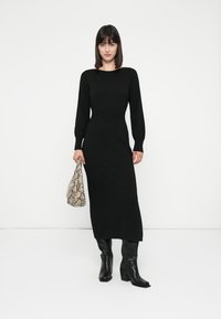 Abito nero a maniche lunghe in maglia con silhouette aderente, scollatura rotonda e lunghezza fino al polpaccio. Abbinato a una borsa beige a motivi e stivali neri.