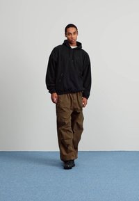 Črna pulover s kapuco z sprednjim žepom in nastavljivimi vrvicami, v kombinaciji z ohlapnimi rjavimi cargo hlačami. Črne čevlje dopolnjujejo videz.