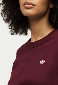 Close-up of a person with curly hair wearing a burgundy sweatshirt featuring a small white Adidas logo on the chest.
