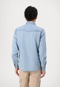 Camisa de mezclilla azul claro con cuello puntiagudo, mangas largas y puños abotonados. Presenta una tela sutilmente texturizada y un dobladillo curvado.