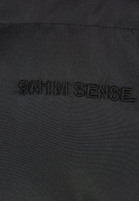 Țesătură neagră cu text brodat "9N1M SENSE". Textura pare netedă, având un finisaj mat și fără modele sau accente vizibile.