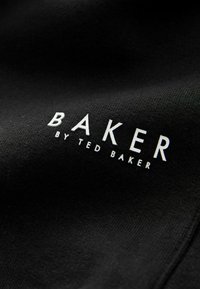 Čierna tkanina s textom "BAKER by TED BAKER" v bielej, čistej sans-serif písme, umiestnená výrazne po celej ploche.
