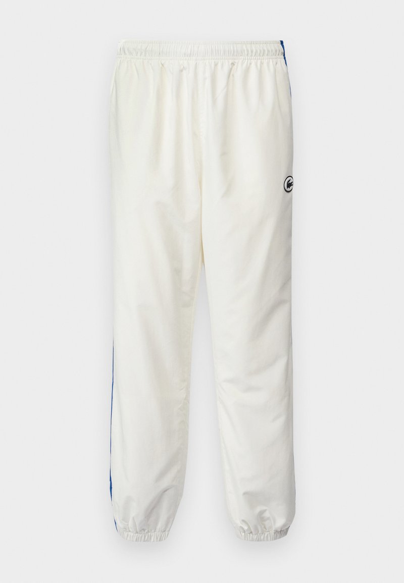 Pantalons de sport blancs avec taille élastique, comportant une bande latérale bleue et un petit logo noir sur la hanche gauche. Tissu extensible et léger.