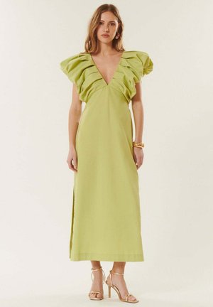 Femme portant une longue robe vert citron avec des épaules volantées, un décolleté en V profond et des sandales dorées à talons hauts, debout devant un fond uni.