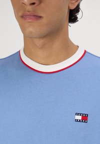 Camiseta de algodón azul claro con cuello redondo blanco y rojo, que presenta un pequeño parche del logo de Tommy Jeans en el lado izquierdo del pecho.