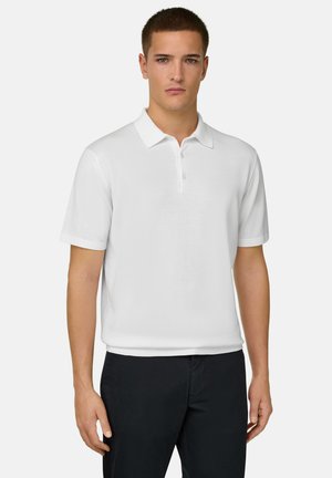 Giovane uomo che indossa una polo bianca semplice a maniche corte e pantaloni neri, in piedi davanti a uno sfondo grigio chiaro.