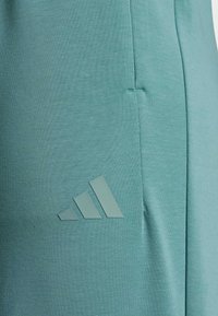 Türkise Sporthose aus strukturiertem Material, ausgestattet mit einer Seitentasche und einem erhabenen Adidas-Logo in passendem Farbton.