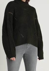 Pull en maille vert foncé avec un col montant, des épaules tombantes et des détails en point ouvert. Il présente une coupe décontractée et des poignets et ourlet côtelés.