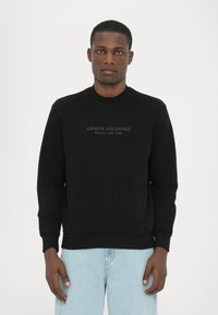 Juodas džemperis su apvalia iškirpte, minkšta tekstūra ir siuvinėtu "Armani Exchange Milano/New York" logotipu krūtinėje.