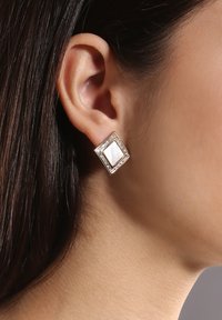 Quadratische Ohrringe mit einem Mittelstück aus Perlmutt, umgeben von einem goldfarbenen Rahmen, der mit kleinen, klaren Schmucksteinen verziert ist.