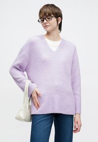 Vijoličen pulover z V-izrezom iz mehke pletenine, z rebrastimi manšetami in spodnjim robom. Kombiniran z modrimi kavbojkami in majhno belo torbico.