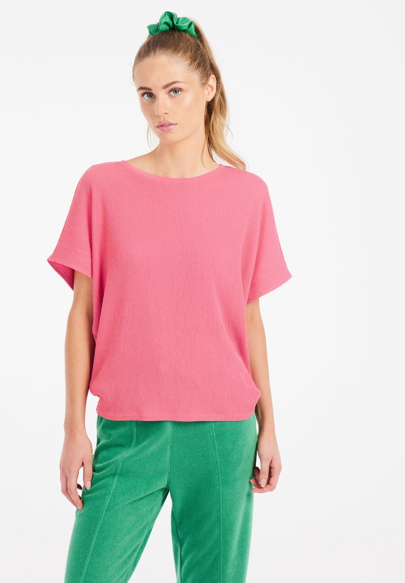 Top in maglia a coste rosa con vestibilità ampia e maniche corte larghe, abbinato a pantaloni verdi testurizzati. Colori solidi, design casual.