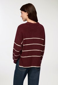 Kobieta z ciemnobrązowymi włosami, ubrana w bordowy sweter w białe poziome paski i niebieskie dżinsy, stojąca tyłem na białym tle.
