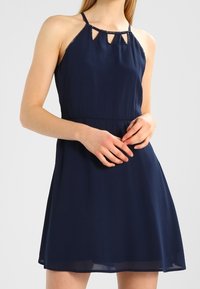 Kvinna som bär en ärmlös marinblå klänning med nyckelhålsskärningar vid halsringningen och tunna axelband, stående med händerna ihopknäppta.