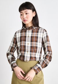 Bruin en crèmewit geruit overhemd met hoge kraag, voorzien van ruches aan de manchetten en metallic knopen. Gemaakt met een effen groene rok.
