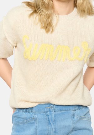 Femme portant un pull en maille crème avec un texte jaune "summer" et un jean taille haute bleu clair, les mains sur les hanches.
