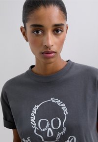 Tricou gri din bumbac cu mâneci scurte și decolteu rotund, având un desen alb cu craniu și inscripții. Textură netedă, potrivire casual.