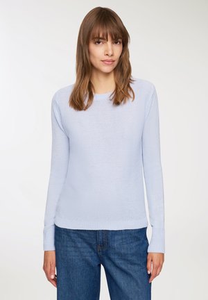 Kobieta z prostymi brązowymi włosami, ubrana w jasnoniebieski sweter dzianinowy i niebieskie dżinsy, stojąca na tle gładkiej białej ściany.