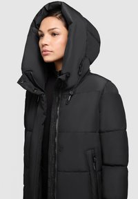 Jachetă puffer neagră cu un glugă mare, design matlasat, fermoar frontal și buzunare laterale cu accesorii decorative din metal.