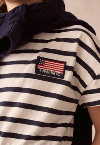 Randig bomull t-shirt med marina och vita horisontella ränder, prydd med ett röd, vit och blått märke som visar en amerikansk flagga och "SUPERDRY."