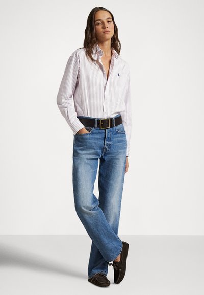 Sieviete, kas valkā gaiši rozā svītrainu krekla kreklu, ielikta kokvilnas džinsās ar melnu jostu un tumši brūnām mokasīnu kurpēm, stāv, vienu roku ieliekot kabatā.