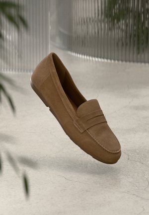 Zapato de ante marrón tipo mocasín con punta redondeada, costuras decorativas y una banda en el empeine. Textura suave y suela flexible.